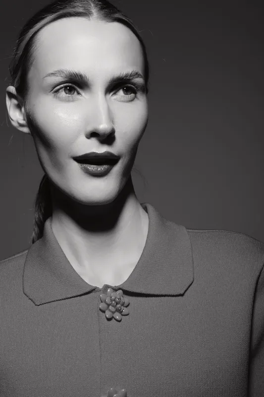 Portrait of a young white woman with light skin and slicked-back hair wearing a textured shirt with decorative buttons in black and white. Studio lighting highlights facial features.