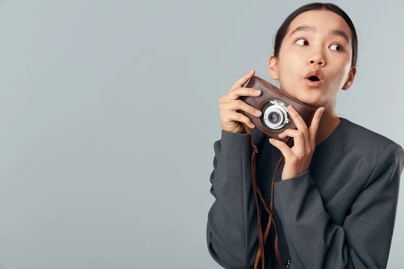 Young woman holding vintage camera close to her face with surprised expression, wearing dark sweater and looking sideways on gray background in studio portrait.