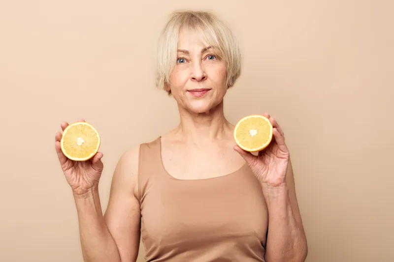 Mature woman with short blonde hair holds two halves of fresh orange fruit in hands against beige background. Concept of healthy lifestyle, natural vitamins, skincare, wellness, and nutrition.