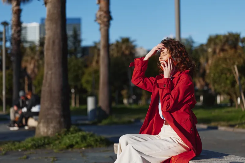 Young woman with curly hair wearing a red jacket talks on a smartphone while sitting on a bench in a sunny park with palm trees in the background.