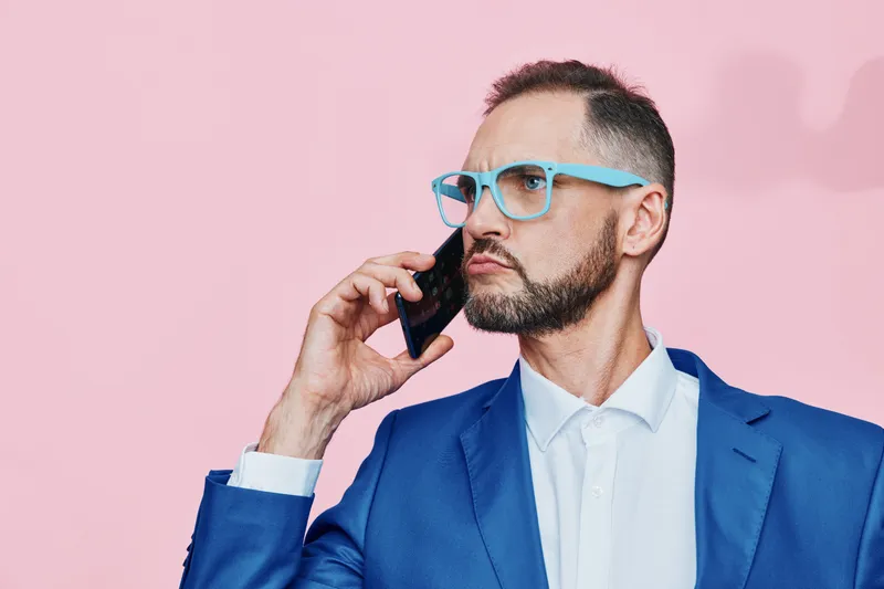 A professional man in a blue suit on a solid pink background, conveys focus and confidence while he speaks on a phone, presenting a strong business presence and clear communication.