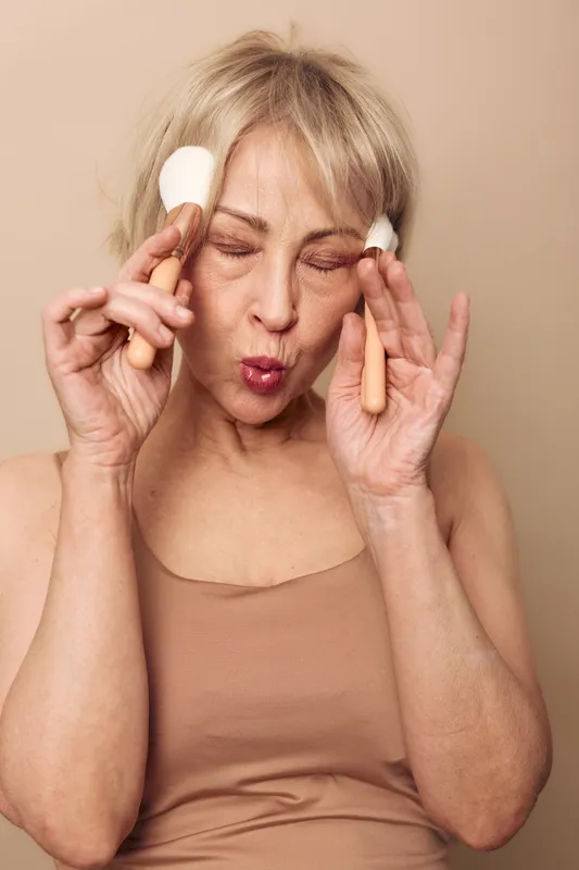 Blonde mature woman in beige top applies makeup brushes on face with eyes closed, playful expression. Studio shot on neutral background. Concept of beauty routine, self-care, and aging gracefully.