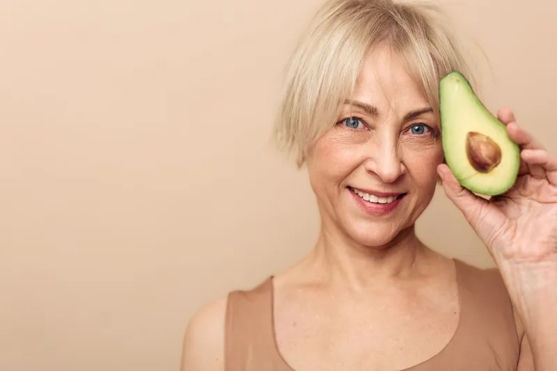 Smiling mature woman with short blonde hair holds fresh avocado half near face. Natural skincare and healthy lifestyle concept with organic fruit on beige background in soft lighting.