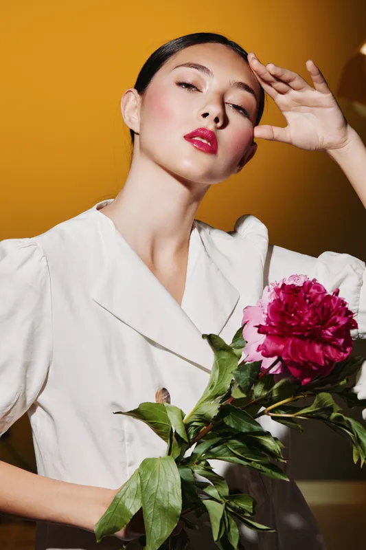 Young woman with glossy red lips and minimalistic makeup poses holding a vibrant pink flower. She wears a white structured blazer against a warm orange background. Beauty creative fashion concept.