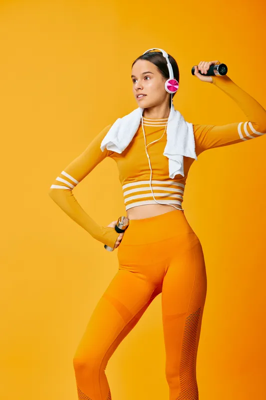 Fitness woman in bright orange athletic outfit poses in a dynamic workout stance, towel draped over shoulders and wireless headphones on, vibrant orange studio backdrop.