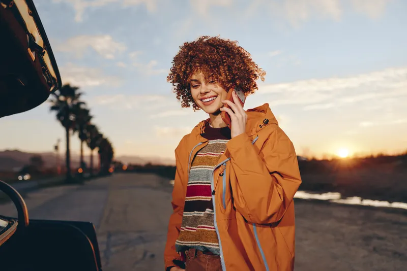 Woman with curly hair smiling at sunset outdoors on a road, wearing jacket, standing by a car, casual travel lifestyle, warm evening light, relaxed mood