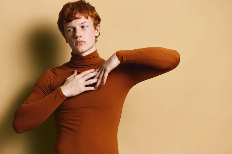 Young man with red hair wearing brown turtleneck posing with hand on chest and elbow raised against beige background with soft shadow lighting.