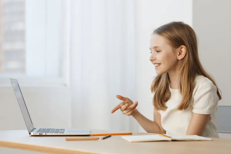 Cheerful young girl engaging in a video call, smiling and gesturing, wearing a casual white t shirt, in a bright, minimalistic room with natural light filtering in