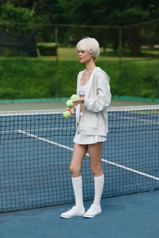 Young female tennis player in a sporty outfit holding tennis balls, expressing confidence on a tennis court, surrounded by greenery, ideal for sports themed content