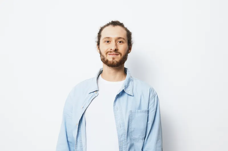 Smiling young man with light brown hair and beard wearing a casual blue shirt posing against a white background, radiating positivity and confidence