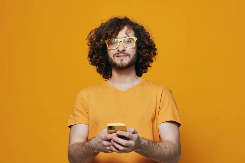 Young man with curly hair in yellow glasses holding a smartphone against a bright orange background, showcasing a cheerful and modern lifestyle