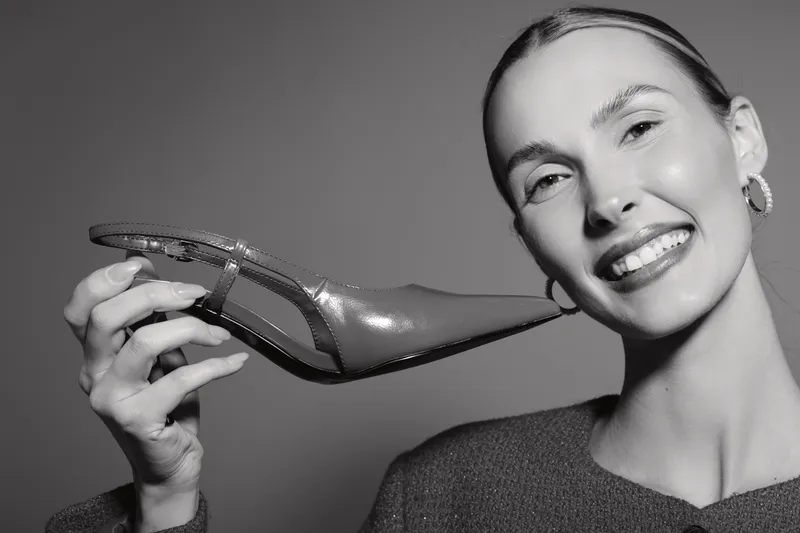 Smiling Caucasian woman with fair skin holds high heel shoe near face, studio portrait on grey background. Concept of fashion accessory and beauty.