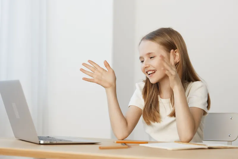 Happy young girl waving at her laptop during an online call, wearing a casual white shirt, in a bright, minimalistic room, capturing joy and connection