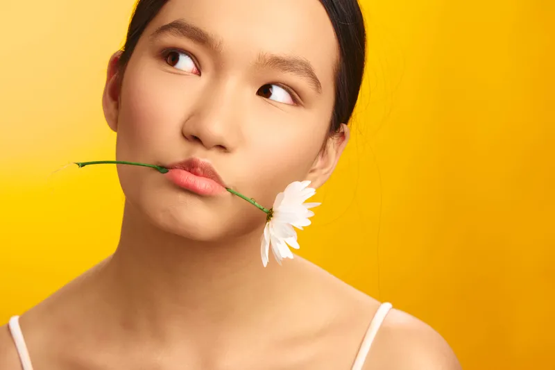 asian woman with a delicate expression gazes softly to the side while holding a white flower between her lips, creating a serene portrait against a warm yellow background.