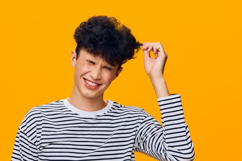 young man with curly hair wearing striped shirt laughs with eyes closed against bright orange background, expressing joy and playfulness, casual style portrait