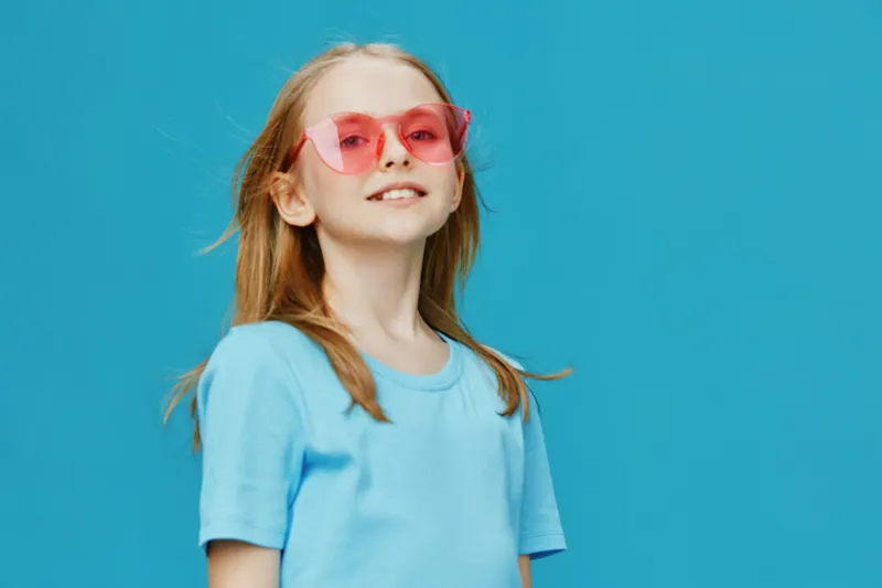 Cheerful girl wearing pink sunglasses and a blue t shirt against a vibrant turquoise background, showcasing a playful summer vibe and youthful joy