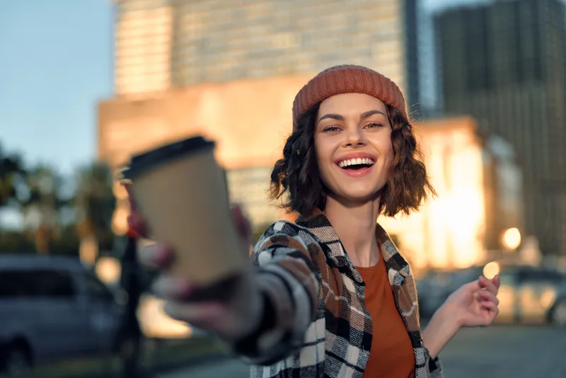Woman smile coffee urban beanie candid - joyful young woman offering takeaway cup in golden hour glow, authenticity and mindful living captured in emotional storytelling portrait.