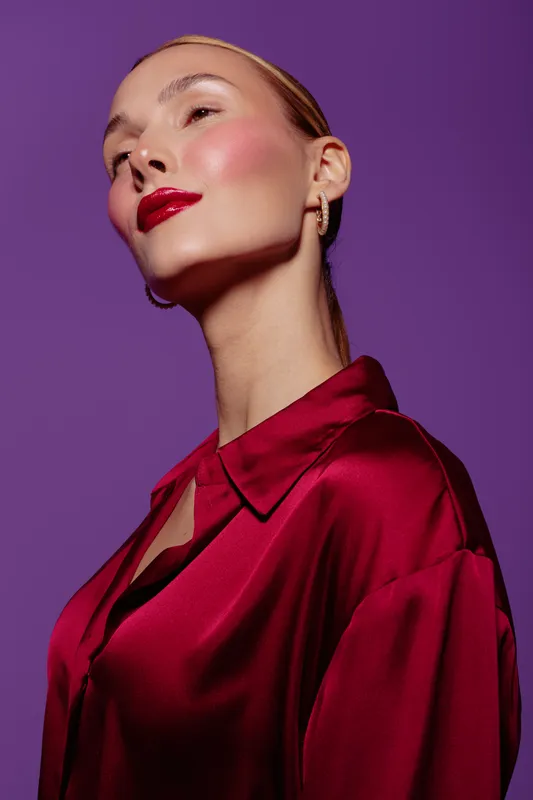 Elegant woman with red lipstick and shiny satin blouse poses confidently against purple background. Concept of fashion, beauty, glamour, and modern style portrait.