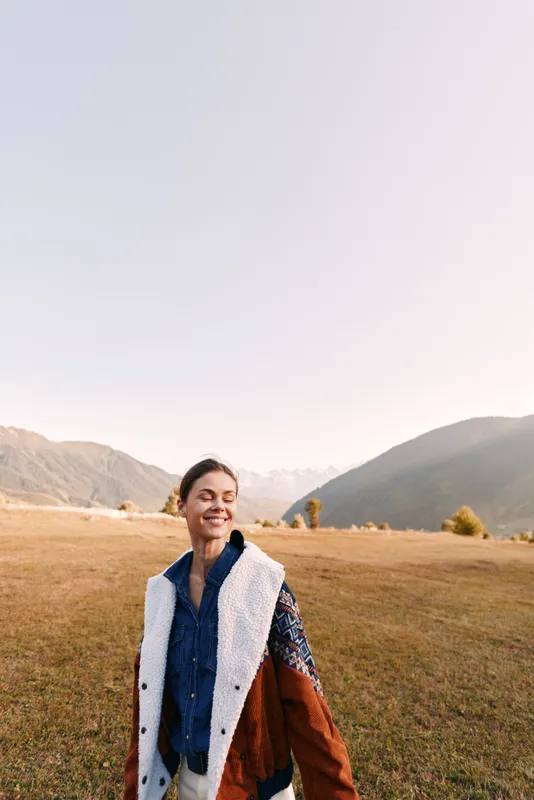 Woman smiling in a field with mountains in the background, wearing a warm coat in autumn. Outdoor portrait shows serene landscape, casual fashion and peaceful nature for travel mood.