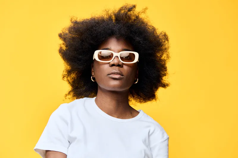 Stylish young woman with curly hair wearing sunglasses, posed against a vibrant yellow background, exuding confidence and modern fashion