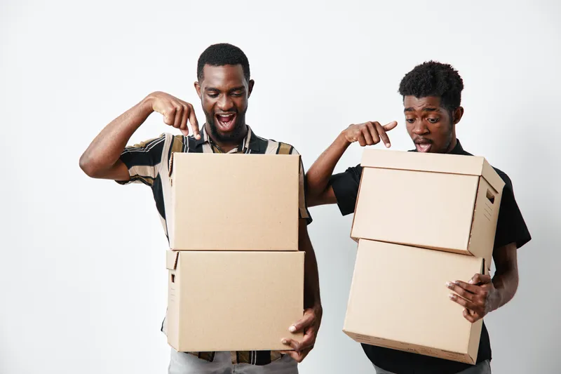 Two young black men holding cardboard boxes and pointing at them with excitement on white background. African ethnicity, dark skin, casual clothing and happy expressions.