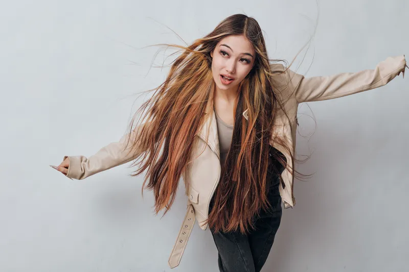 Young woman with long hair wearing beige jacket and black pants posing with arms spread on gray background. Casual style, motion and expressive look for fashion or lifestyle concept.