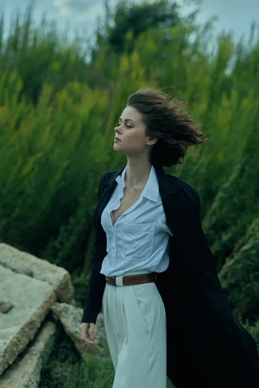 Woman in white shirt and dark coat billowing in the breeze beside rocks and green landscape, showcasing outdoor fashion, movement and natural elegance.