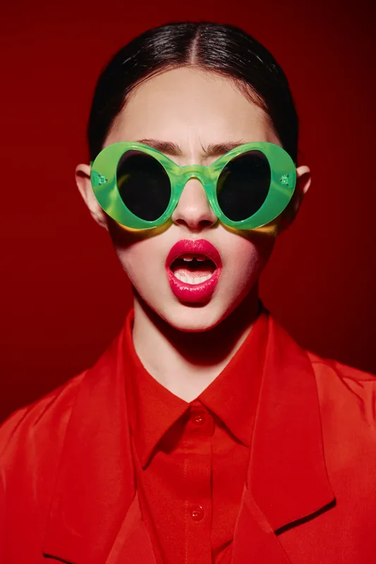 Fashionable young woman wears oversized green sunglasses and bright red outfit on vibrant red backdrop. Her open mouth and bold makeup express cool attitude and beauty concept.
