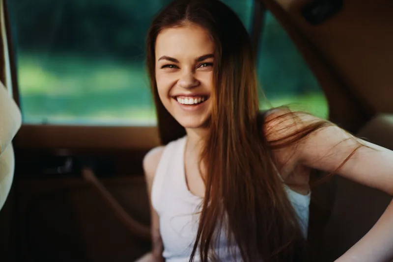 Young woman smiling joyfully in a car, casual outfit and long hair, capturing a moment of happiness and freedom in nature's backdrop.