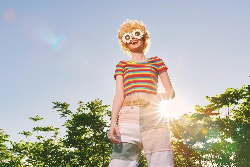 Smiling woman wearing a striped crop top and flower mask stands outdoors under bright sunlight, enjoying a carefree summer moment. Retro color tones and nature backdrop convey lifestyle, joy, and