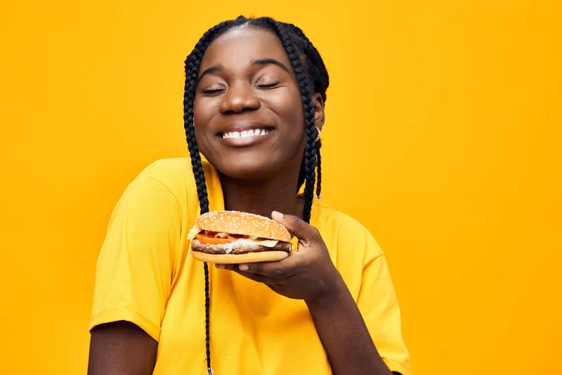 Joyful young Black woman with braids enjoying a delicious burger against a vibrant yellow background, showcasing happiness and healthy eating
