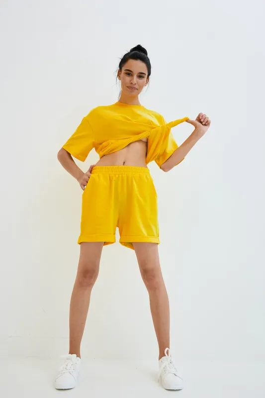 young woman in yellow casual outfit posing confidently against white background, wearing shorts and t-shirt with white sneakers, sporty and modern style