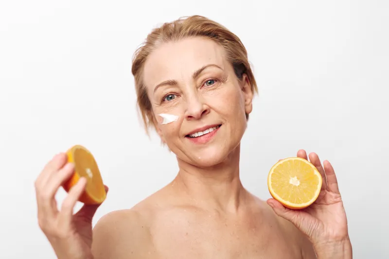 Smiling mature woman with short hair holding two halves of orange fruit near face, applying natural skincare cream on cheek. Concept of healthy lifestyle, beauty, and organic self-care.