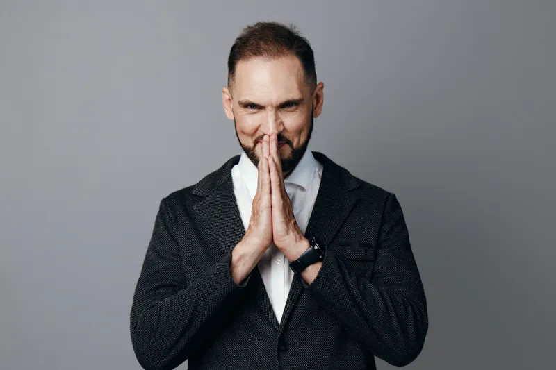 A professional man in business attire stands against a solid colored background, projecting focus and determination as hands are pressed together in a thoughtful pose.