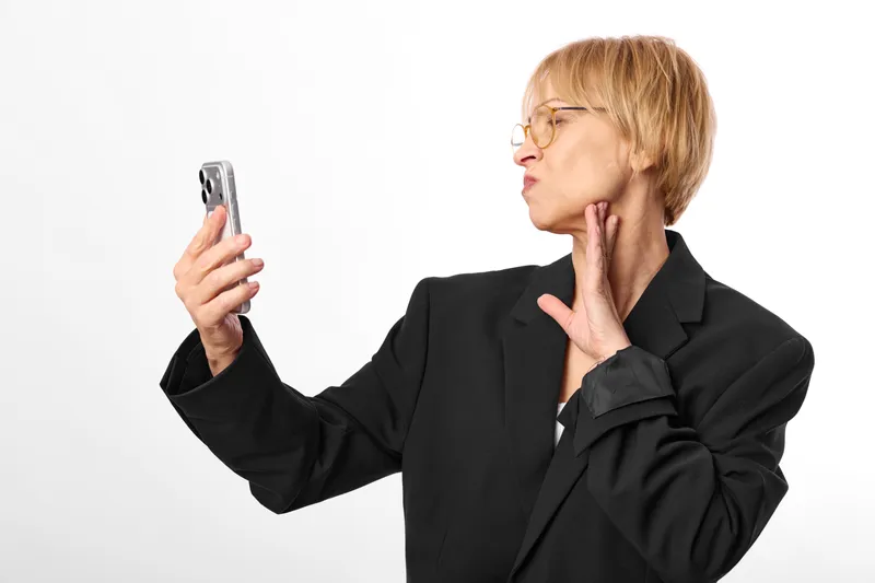Elderly businesswoman with glasses looks surprised, holding a smartphone and touching her cheek, worried about a message, white studio background, anxious reaction