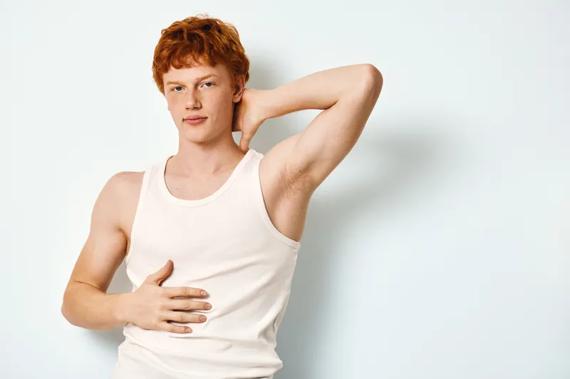 Young man with red hair in white tank top posing with hand behind head and other on stomach, studio portrait with plain light background and confident expression.