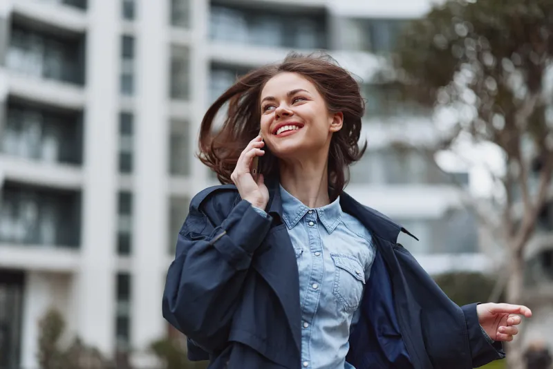 A cityscape unfolds as a smiling woman in a denim shirt and blue coat talks on a phone, wind tousling her hair in an active urban moment.