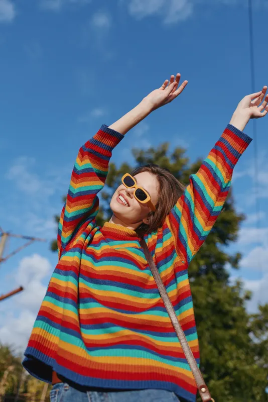 Young woman in her twenties wearing a rainbow sweater and yellow sunglasses, smiling outdoors in an urban park with arms raised, casual outfit, joyful lifestyle and sunny mood.