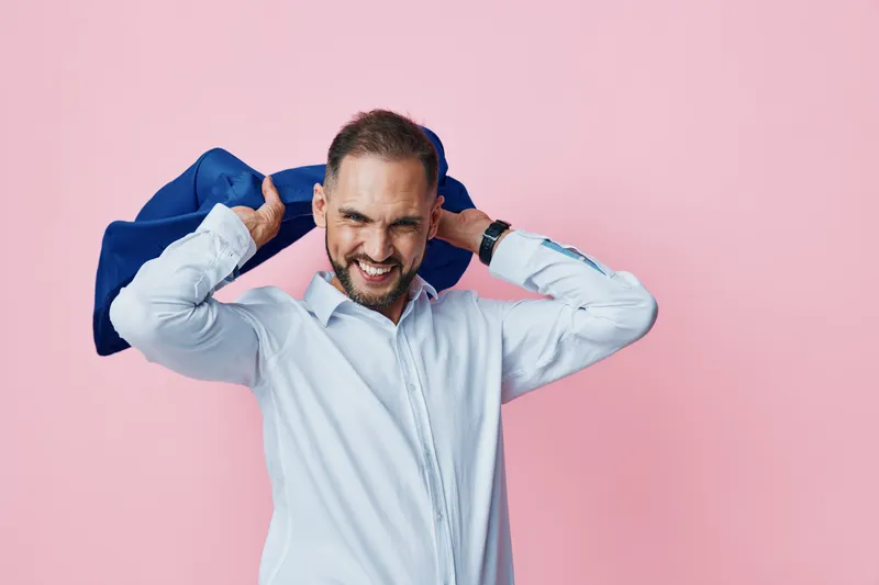 A confident business professional stands against a solid pink background, pulling on a jacket with a playful smile, illustrating dynamic mood and readiness.