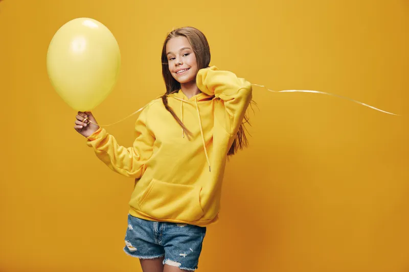 Smiling girl in yellow hoodie holding a yellow balloon with braided hair standing against yellow background. Happy child enjoying playful moment with casual style and confident pose.