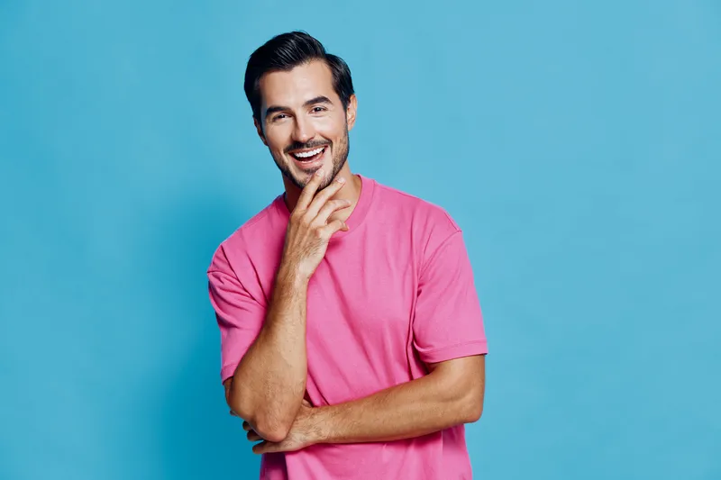 Smiling happy cheerful young man wearing pink t-shirt looking at camera with hand on chin isolated on bright blue background. Joyful lifestyle concept.