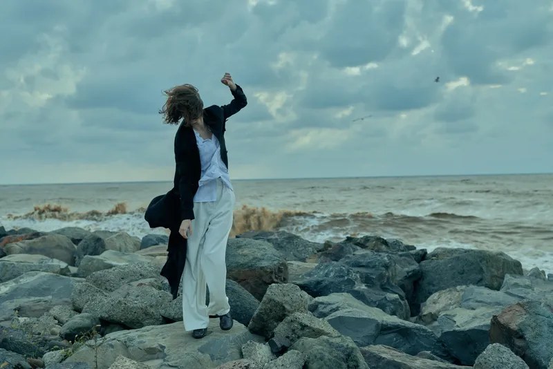 Figure on rocky shoreline in windy weather, flowing white trousers and dark coat, poised mid step among spray and rugged stones by the sea