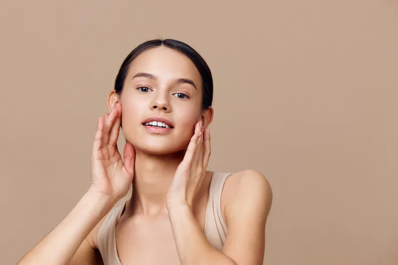 Young woman with smooth skin expressing joy, wearing a nude top against a neutral beige background, emphasizing beauty and self care in a minimalist style