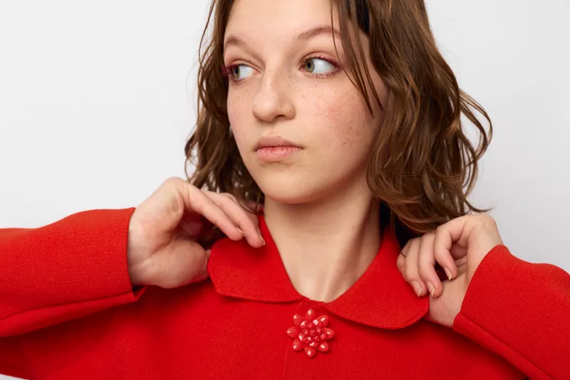 Young woman in red sweater adjusts collar while looking sideways. Casual style, natural makeup, and soft lighting create a calm and thoughtful mood in studio setting.