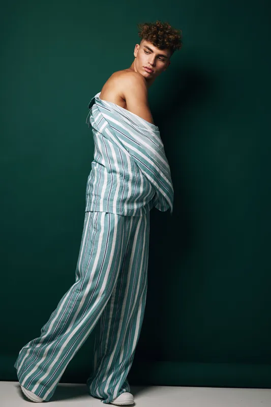 Young man posing in striped pajamas with bare shoulder against dark background, fashion portrait with casual style and relaxed mood in studio lighting setup.
