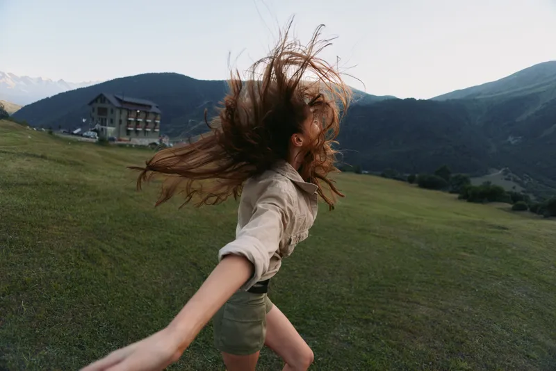 A young adult woman with windblown hair runs across a green hillside, arms extended in a free, joyful moment against a backdrop of mountains and open sky.