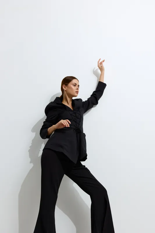 Fashion woman in black stylish outfit poses confidently against white wall, showcasing modern minimalistic look with high contrast and sharp shadows, highlighting contemporary style.