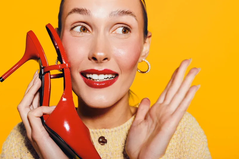 Young woman with red lipstick holds red high heel shoe near face, smiling and looking aside on bright yellow background. Concept of fashion, beauty, style, and happiness.