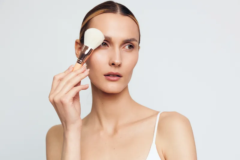 Young woman with blonde hair applies powder to her face using a large makeup brush while looking slightly to the side against a plain light background.