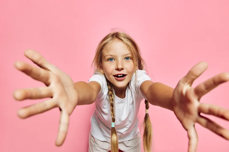 Happy young girl with braided hair dressed in white reaching arms forward joyfully on a pastel pink background. Studio shot with playful and energetic mood.
